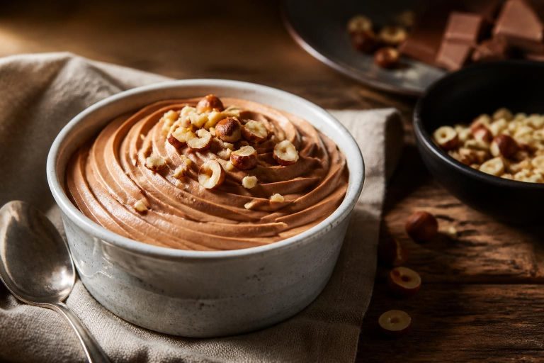 Bol de crème mousseline à la pralinoise onctueuse, avec une tablette de pralinoise et des noisettes caramélisées posées sur un plan de travail en marbre blanc